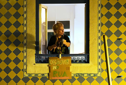 A woman bangs on a pot lid from a window as demonstrators gather to demand affordable housing outside Portugal's Parliament in Lisbon, Tuesday night, Oct. 27, 2026. The sign reads in Portuguese: "Out with the (mis)government." 
