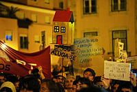 In Photos | Portugal’s Housing Crisis Fuels Protests as Residents Push Back Against Soaring Rents | Photo: AP/Armando Franca : A protester carries a cardboard house with a "For Rent" sign during a demonstration demanding affordable housing outside Portugal's Parliament in Lisbon, Tuesday night, Oct. 27, 2026.