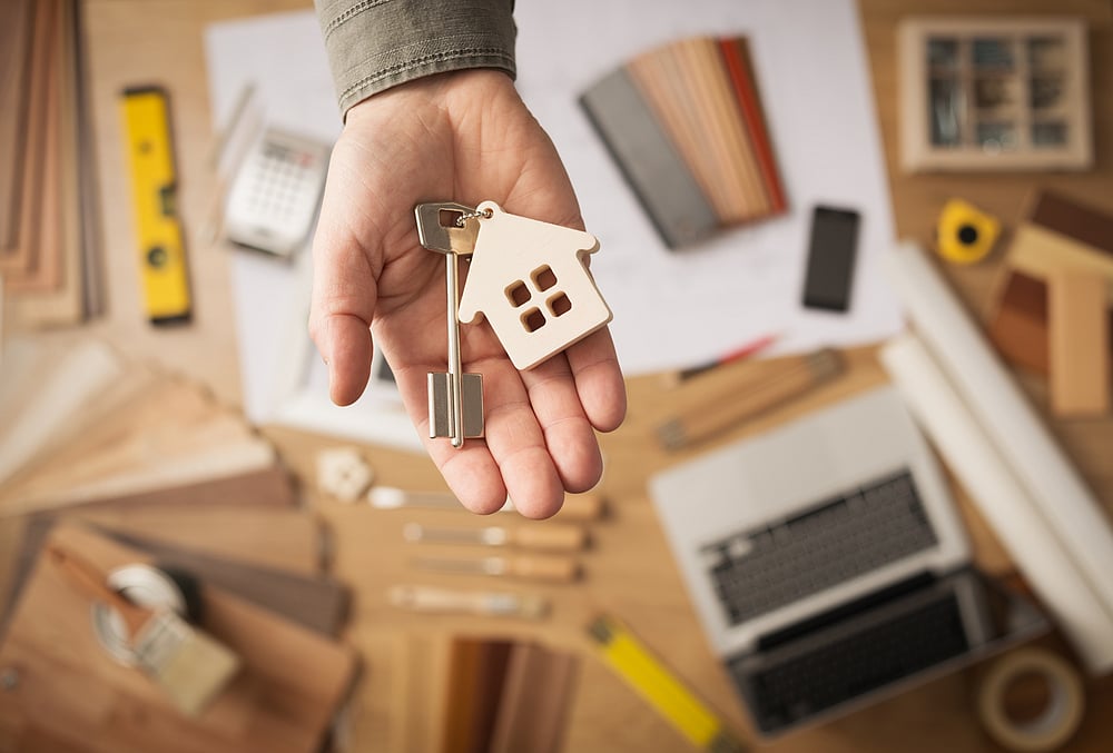 Hand holding house-shaped keychain with a key, background of laptop, tools, and home design samples