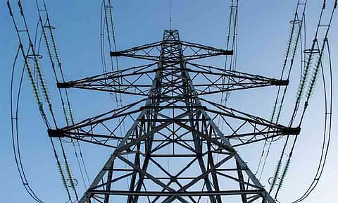 A high-voltage electricity transmission tower stands against a clear blue sky in this undated photograph. The steel lattice pylon serves as critical grid infrastructure, using suspended strings of glass or ceramic disc insulators to carry overhead power lines that distribute electricity across long distances.
