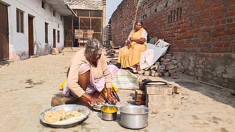 An elderly woman sat on the ground, engrossed in cooking on a stove. Nearby, was another woman, holding a stick, perched on a pile of bricks. 