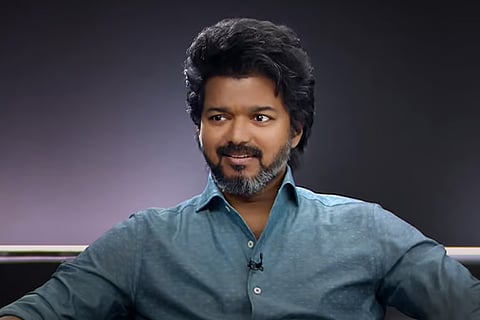 Actor Vijay in a blue shirt, seated in front of a grey background. 