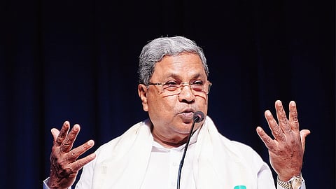 Karnataka Chief Minister Siddaramaiah speaking into a microphone at a podium. He is wearing a white shirt and white shawl and has spectacles. Both his arms are raised in a gesture mid-speech