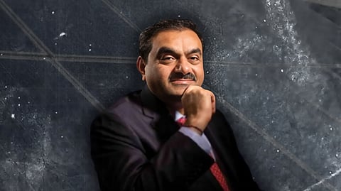 Adani group chairperson Gautam Adani, wearing a dark suit and red tie is posing with a thoughtful expression, resting their chin on their hand. The background features a textured, abstract gray pattern.