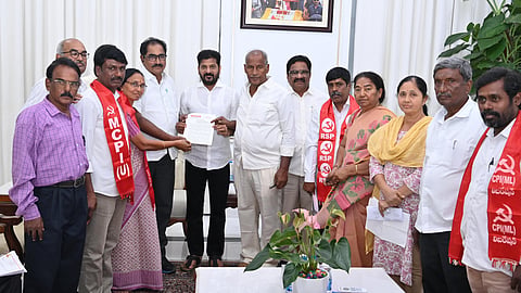 A group of people, some wearing red scarves with Left political party logos, gather around Telangana Chief Minister Revanth Reddy holding a document. They are standing in a room with a table, some papers, and a potted plant. A framed picture hangs on the wall behind them.
