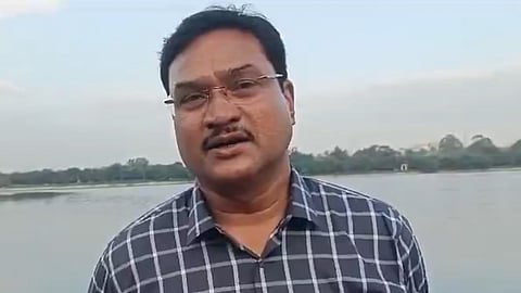 AV Ranganath, Commissioner of Hyderabad Disaster Response and Asset Protection Agency (HYDRAA), wearing glasses and a blue checked shirt stands in front of a calm lake, with trees and a clear sky in the background.
