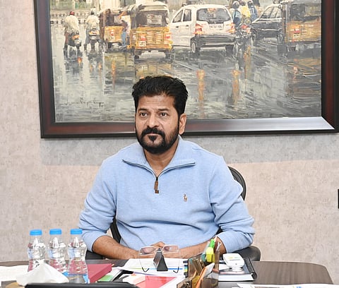 Telangana Chief Minister A Revanth Reddyin a blue full sleeved t shirt staring ahead of him. There's a painting of a road with vehicles in rain behind him. There are three water bottles and stationery on the table in front of him. 