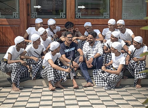 A group of people sits together in front of a wooden building entrance. Most wear white shirts, checkered pants, and headscarves, holding sticks, likely part of a traditional performance. Two men in modern clothing sit in the centre.