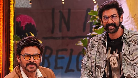 Actor Venkatesh is sitting on a couch while his nephew Rana Daggubati is leaning on the couch standing next to him. They are on a studio set. Venkatesh is wearing a white shirt, white pancha and brown jacket. Rana is wearing a black t shirt with Bob Marley's image and and a black and white overshirt, and a pancha. They are both wearing sunglasses and smiling at the camera. 