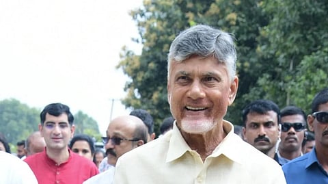 Andhra Pradesh Chief Minister Chandrababu Naidu dressed in a cream-colored shirt and traditional white attire is smiling while walking outdoors. He is surrounded by a group of people, some in casual traditional attire and others in formal security uniforms. Trees and greenery are visible in the background, suggesting a public or cultural event.







