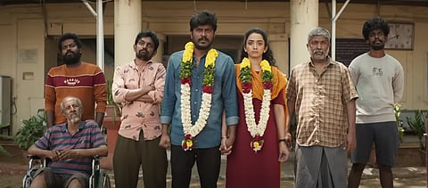 A scene from Kudumbastha: A group of seven stands outside a building. Naveen and Vennila in the center, adorned with flower garlands and holding hands, flanked by five  men. One of them is an elderly man who sits in a wheelchair on the far left.
