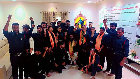 Men and women in black shirts, black pants, and a saffron scarf around their necks stand in a room facing the camera. One person sitting in the middle. Some of them have their fists raised as though raising a slogan. 