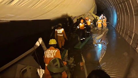 Rescue workers from the National Disaster Response Force (NDRF) of India are seen inside a partially collapsed tunnel. They are wearing orange uniforms and safety gear, including helmets and reflective vests, while working in a muddy and waterlogged environment. The tunnel lining appears damaged, with a large tarp or covering hanging from the ceiling. The team is using stretchers or boards to navigate the unstable ground, and artificial lighting illuminates the scene as they proceed with the rescue operation.