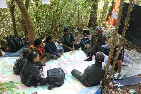 Two civil society members and a group of Maoists dressed in combat fatigues sit in a circle surrounded by trees in a forest. 