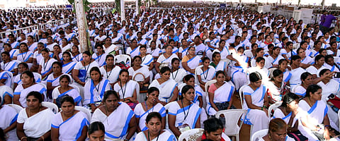 The image shows a large gathering of women wearing white sarees with blue borders, seated in an outdoor setting. Many of them have ID cards around their necks, suggesting they might be part of an organized group or workforce. The crowd appears attentive, possibly attending a conference, seminar, or a government-related event. The women in the image are ASHA (Accredited Social Health Activist) workers. 