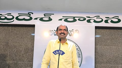 Vizianagaram TDP MP Kalisetti Appala Naidu wearing a yellow shirt is standing at a podium with a microphone, smiling. The backdrop behind him has text in Telugu and a circular emblem, suggesting an official or political setting. The podium has a label that reads "I&PR," indicating an Information and Public Relations setting.