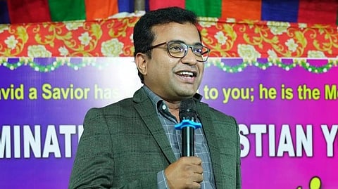 A man wearing glasses and a dark blazer is speaking into a microphone with a bright expression. He stands in front of a colorful backdrop with Christian-themed text and decorations. The background features a mix of purple, yellow, and red colors with floral designs.








