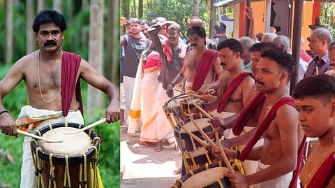 Traditionally dressed Sakkeer Hussain playing a chenda, a cylindrical percussion instrument, in an outdoor setting with lush green trees in the background. He is wearing a white dhoti with a colored border, a maroon cloth draped over one shoulder, and a gold chain around his neck.
