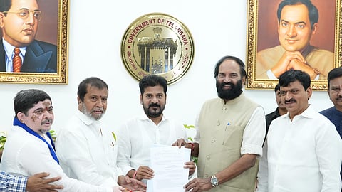 A group of Indian politicians pose together holding a document in front of the Telangana Government emblem. Behind them are portraits of BR Ambedkar and Rajiv Gandhi. The central figure, Revanth Reddy, is flanked by others including Mallu Bhatti Vikramarka, all dressed in formal Indian attire, some in white and one in a beige vest.