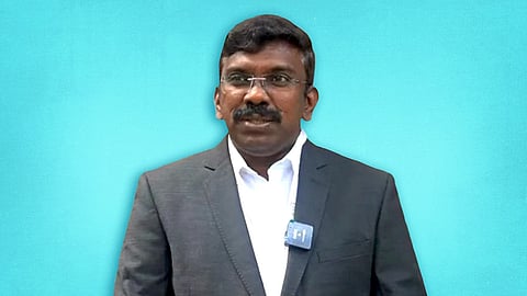 An image of Reny Wilfred in a black blazer and white shirt. The image has a blue background