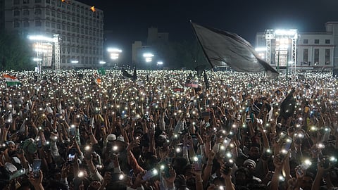 A massive nighttime protest with thousands of people holding up their lit mobile phones, creating a sea of lights. The crowd is densely packed, and some participants wave flags and hold signs. Large stage lights illuminate the area, and prominent buildings are visible in the background.