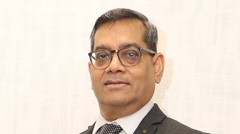 A middle-aged man wearing glasses, a dark grey suit, a white shirt, and a black polka dot tie, standing against a light textured background and looking confidently at the camera.