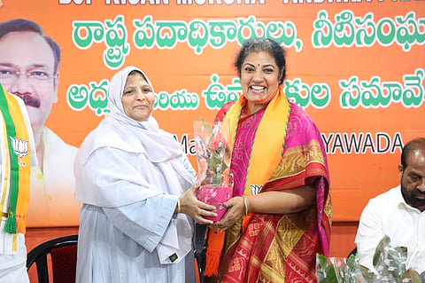 Two women are standing and smiling at a political event. One woman, wearing a white traditional outfit and hijab, is handing a small potted plant to the other woman, who is dressed in a pink and gold saree with a saffron BJP scarf. Behind them is an orange backdrop with Telugu text and the BJP party symbol. A man is seated on the right side of the image.