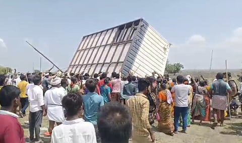 A large crowd of villagers, including men and women, are seen forcefully toppling a metal structure using sticks and sheer force during a protest. The structure appears to be part of an industrial setup. The scene is unfolding in an open area under a clear sky, indicating a heated and collective act of resistance.
