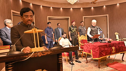 A formal swearing-in ceremony is taking place in an ornate hall with a chandelier and wooden paneling. A man in a black suit speaks at a podium with the Indian national emblem. In the background, another man in a cream-colored vest stands at a microphone reading from a document, taking the oath. Several officials, including security personnel and dignitaries, are present, some seated on red and gold chairs. A table with a red velvet cover bearing the Indian emblem is in front of the man taking the oath.