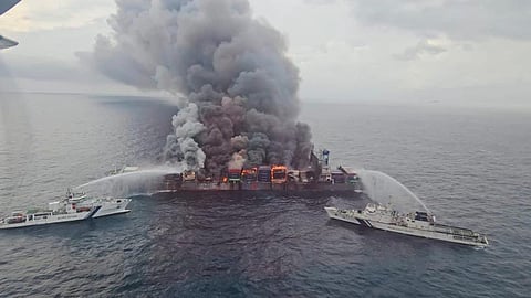 Aerial view of a large container ship engulfed in thick black smoke and flames, with two Coast Guard vessels spraying water to extinguish the fire at sea