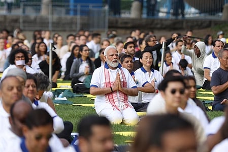 Prime Minister Narendra Modi sitting cross-legged with hands folded in a namaste gesture during a yoga session in a park, surrounded by participants dressed in white.
