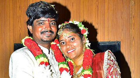 A newlywed couple dressed in traditional South Indian attire poses closely together, smiling. The groom wears a white sherwani and the bride is adorned in a red and cream saree with gold embroidery. Both wear garlands made of red, white, and yellow flowers and have ceremonial decorations like bangles, gold jewelry, and floral headpieces. They are holding hands affectionately against a wooden-panel background.