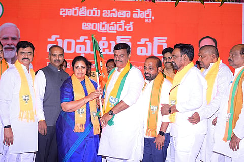 A group of Bharatiya Janata Party (BJP) leaders and members are seen on stage at a political event in Andhra Pradesh. A woman in a blue saree, wearing a BJP scarf, hands over a party flag to a man in white traditional attire, also wearing a BJP scarf. Other leaders stand alongside them, all dressed in white with orange BJP scarves, against a bright orange backdrop displaying the party's name in Telugu. Portraits of national BJP leaders, including Prime Minister Narendra Modi, are partially visible in the background. The atmosphere appears celebratory and symbolic of new membership or alliance.