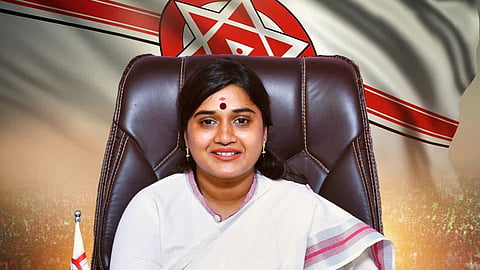A woman dressed in a white saree with a pink border is seated at a wooden desk, hands clasped, smiling confidently. She has a red bindi on her forehead and is wearing gold bangles and earrings. Behind her is a large flag featuring the Janasena Party symbol—red and white stripes with a red circle and white star in the center. A small Janasena flag is placed on the desk to her left, and a neat stack of files is on her right. The background includes a large, cheering crowd, adding to the political tone of the image.