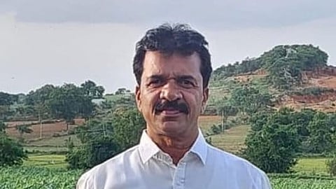 A man wearing a white shirt and white pants stands in a lush green field, likely a farm with young crops. He has short black hair and a mustache, and he is facing the camera with a neutral expression. In the background, there are trees, hills with red soil patches, and a partly cloudy sky.