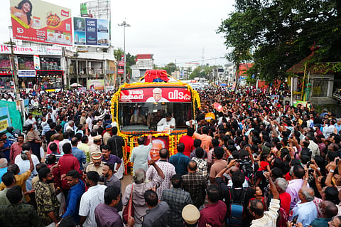 A large crowd gathers on both sides of a street in Kerala as a flower-decorated bus carrying the body of V S Achuthanandan moves slowly during his funeral procession, with people taking photos, holding banners, and police managing the crowd.