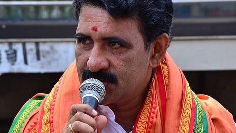 B Gopalakrishnan holding a wired microphone. Red religious mark on his forehead. Saffron scarves on his shoulder