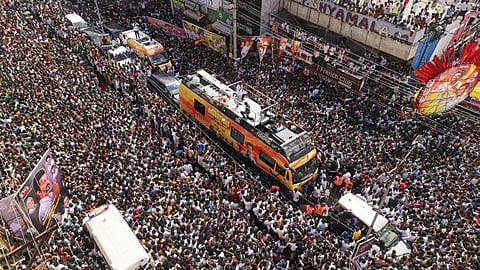 An extremely crowded aerial photo shows a large political rally or procession in an urban area. A brightly colored, custom-made campaign bus, predominantly yellow and orange, moves slowly through the center, surrounded by an immense throng of people, many of whom are wearing white. The bus has a platform on top where several people are standing. On the right, signs, banners, and building facades are visible, including one with the text "French Club." Large posters, likely featuring a politician, are also displayed among the crowd. The density of the crowd suggests a major political event in India.
