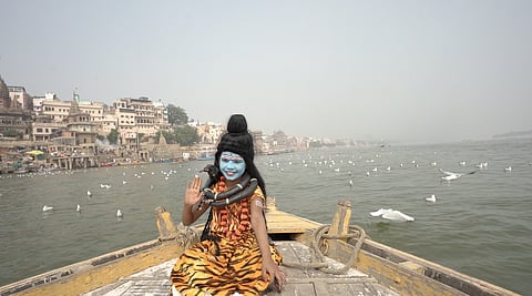 A low-angle, wide shot shows a young child in the attire of the Hindu deity Lord Shiva sitting on the bow of a wooden boat on a river, likely the Ganges (Ganga), in Varanasi, India.

The child, facing the camera, has their face painted blue with white markings (similar to Shiva's third eye), wears a high black wig styled into a bun, and has a garland of plastic black "snakes" around their neck, one of which rests on their shoulder. They are wearing a brightly patterned, orange and black tiger-print cloth, suggesting the traditional depiction of Shiva's tiger skin. The child is raising their right hand in a wave.

In the background, the river is wide and murky, with many white seabirds flying or floating on the water. On the left bank, the dense, ancient stone and stucco buildings of a ghat in Varanasi rise up. The sky is a pale, hazy white. The foreground of the wooden boat is dominated by the child and the worn, weathered wood of the vessel.