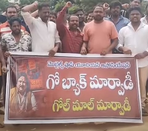 A group of men is holding a large red and white banner in what appears to be an outdoor protest. The men are mostly raising their fists in the air. The banner features text in Telugu, which includes:

'మొబైల్స్ షాప్ యూనియన్ అసోసియేషన్' (Mobiles Shop Union Association)

'గో బ్యాక్ మార్వాడీ' (Go Back Marwadi)

'గోల్ మాల్ మార్వాడీ' (Golmaal Marwadi, which can be interpreted as 'Fraud/Deceptive Marwadi').

There is also a picture of a man on the left side of the banner with small text that includes 'GO BACK MARWADI' in English. The image captures a demonstration against Marwadi traders by a Mobile Shops Union Association.