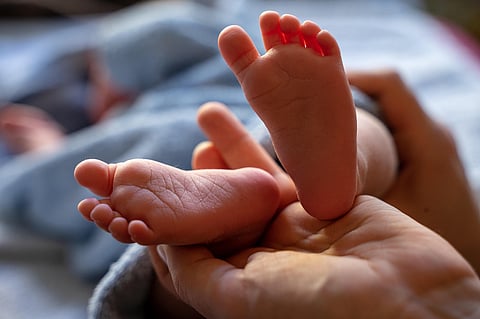 A close-up photograph focuses on a pair of tiny, pink baby feet being gently held in an adult's hands. The baby is lying down, partially covered by a gray blanket visible in the soft-focus background. The lighting is soft and warm, highlighting the texture of the baby's soles and toes.