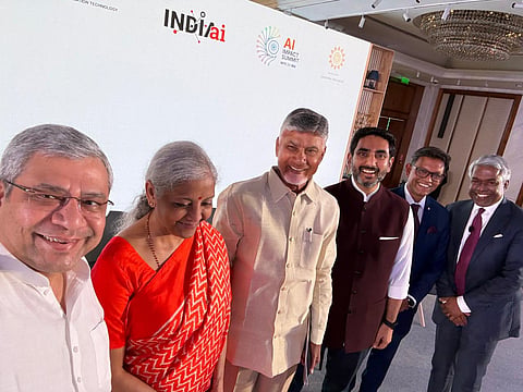 A group photo of six people—four men and two women—standing in a line against a white and wood-paneled backdrop.

From left to right: A man with gray hair and glasses, wearing a white shirt; a woman with gray hair wearing a red and orange sari; a man with light gray hair, wearing a light yellow button-down shirt; a man with dark hair and a beard, wearing a maroon Nehru jacket over a white shirt; a man with dark hair and glasses, wearing a dark blue suit jacket; and a man with gray hair, wearing a dark suit with a red tie.

The backdrop features several logos and text, including "INDIAai", "AI IMPACT SUMMIT 2025", and "ANDHRA PRADESH". A large chandelier is visible in the top right. All six people are smiling at the camera.