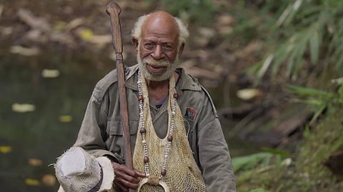 An elderly man wearing a khaki shirt, beaded necklaces, and carrying a walking stick sits near a stream in a lush forest in Papua New Guinea.