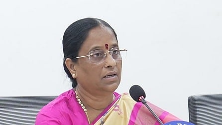 
A clear, medium-close-up image shows a woman, Konda Surekha, seated at a table, speaking into a microphone.

She appears to be middle-aged and is wearing a bright pink blouse with a cream and gold patterned saree. She has a red bindi on her forehead and is wearing glasses. She has gold jewelry, including a necklace and bangles. A stand with the letters "I & PR" (likely Information and Public Relations) is visible near the microphone. She is looking slightly off-camera to her left as she speaks.