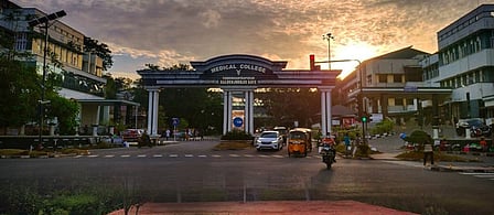 Entrance gate of Thiruvananthapuram Medical College at sunrise, with vehicles and people passing through the road in front of the campus.