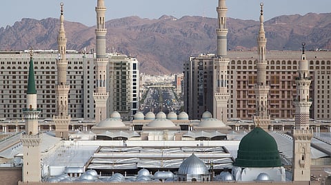 
An eye-level, wide shot captures the immense scale of Al-Masjid an-Nabawi (The Prophet's Mosque) in Medina, Saudi Arabia, against a backdrop of modern high-rise buildings and distant reddish-brown mountains.

In the foreground is the vast, open marble courtyard filled with numerous people. The main structure of the mosque is a complex of beige stone walls featuring numerous arched openings and several large, gray domes.

The most distinctive feature is the Green Dome (Qubbat an-Nabi), located over the resting place of the Prophet Muhammad, which stands prominently on the right side of the mosque complex. To the left of the Green Dome, another taller section of the building features a single slender minaret topped with a green finial.

Rising from the mosque complex are several very tall, pale beige minarets with multiple balconies and pointed tops, dominating the skyline. Behind the mosque, a dense collection of multi-story modern hotels and residential buildings spans the background, with a wide, bustling avenue cutting through the city center. The arid mountains form the final layer of the backdrop.