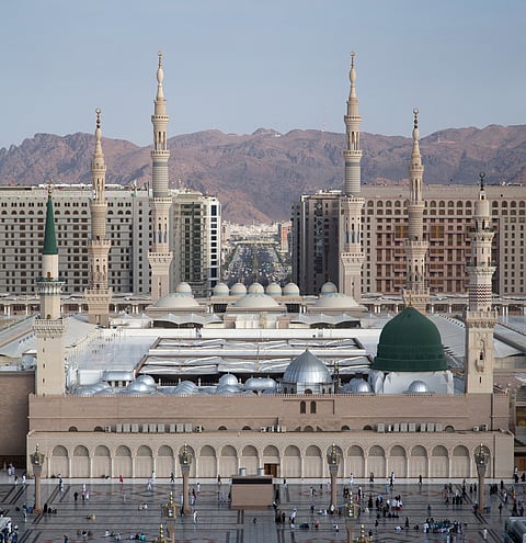 
An eye-level, wide shot captures the immense scale of Al-Masjid an-Nabawi (The Prophet's Mosque) in Medina, Saudi Arabia, against a backdrop of modern high-rise buildings and distant reddish-brown mountains.

In the foreground is the vast, open marble courtyard filled with numerous people. The main structure of the mosque is a complex of beige stone walls featuring numerous arched openings and several large, gray domes.

The most distinctive feature is the Green Dome (Qubbat an-Nabi), located over the resting place of the Prophet Muhammad, which stands prominently on the right side of the mosque complex. To the left of the Green Dome, another taller section of the building features a single slender minaret topped with a green finial.

Rising from the mosque complex are several very tall, pale beige minarets with multiple balconies and pointed tops, dominating the skyline. Behind the mosque, a dense collection of multi-story modern hotels and residential buildings spans the background, with a wide, bustling avenue cutting through the city center. The arid mountains form the final layer of the backdrop.