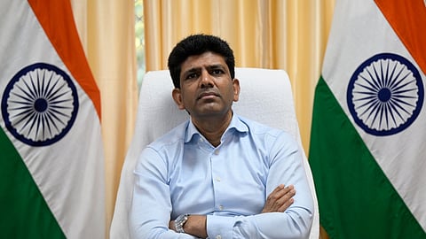 A head-on, medium shot of a man identified as Pemmasani Chandrasekhar sitting in a white chair behind a dark wooden desk.

He is wearing a light blue, long-sleeved collared shirt and a silver wristwatch.

He is looking directly at the camera with a serious expression, his arms crossed on the desk in front of him.

Behind him, on the left and right, are two Indian flags (Tiranga) displayed vertically.

The background features light yellow/cream curtains on either side of a central wall.

A piece of paper and a pen are visible on the desk.