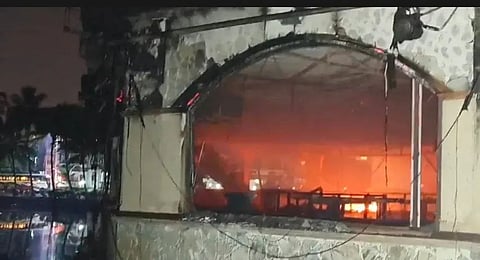 A nighttime photo of a building severely damaged by fire. The arched window frame is blackened and partially melted, exposing the interior where bright orange flames and smoke still burn. Debris and hanging wires can be seen around the opening, and faint reflections of nearby lights appear in water below. Trees and distant structures are dimly visible in the dark background.
