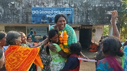 A joyful outdoor scene in front of a Gram Panchayat (village council) office in Amaragiri, Telangana, India. In the center, a tall woman wearing a teal saree and a vibrant orange and yellow marigold garland laughs heartily as she is embraced by two other women.

Around them, several other women in colorful sarees celebrate with broad smiles; one woman on the right has her arm raised in the air in a gesture of victory or cheer. The building in the background features a blue sign with Telugu script that translates to "Gram Panchayat Office, Amaragiri." 